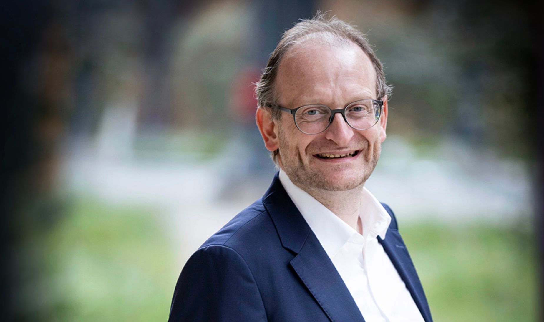 A professional man in a dark blue suit and glasses smiles confidently, ready for a meeting or presentation. A smiling man with glasses and a dark blue suit jacket, blurred background.