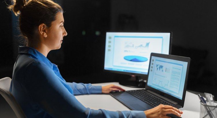 A dedicated professional analyzes data on her laptop and external monitor, working late into the evening with focused concentration. Woman works late at her desk, analyzing data on a laptop and external monitor.