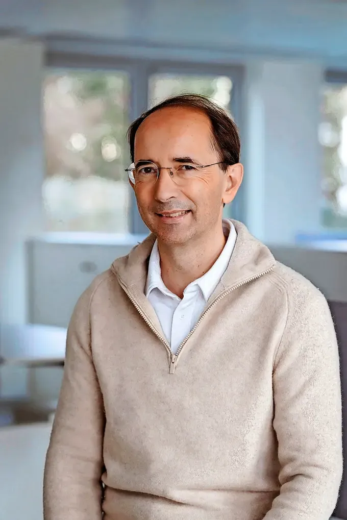 A professional man in a beige sweater and glasses smiles, appearing engaged in an office environment. A professional man with glasses and a beige sweater smiles in an office setting.