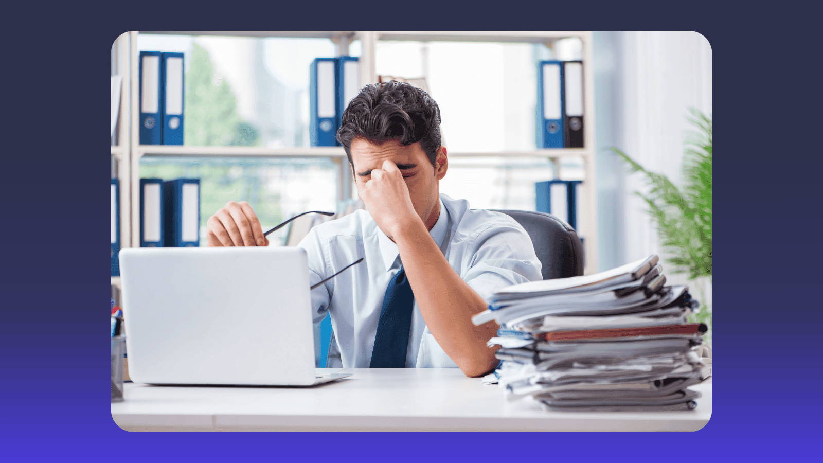 Overwhelmed office worker pinches nose, facing a laptop and stacks of papers.