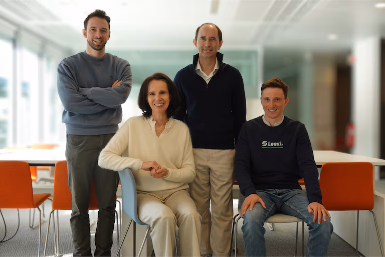 This photo shows four smiling team members, including one wearing a Leexi. shirt, in a bright modern office. Four smiling colleagues, two men standing and two sitting, pose in a modern office.
