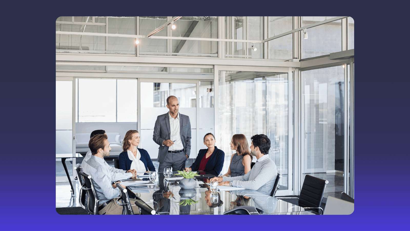 A businessman presents to five colleagues seated around a conference table in a modern office.