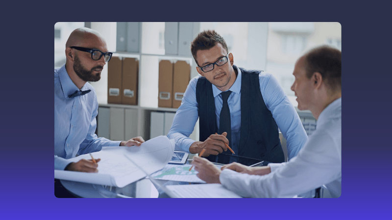 Three businessmen in suits review documents and discuss strategy at an office table.