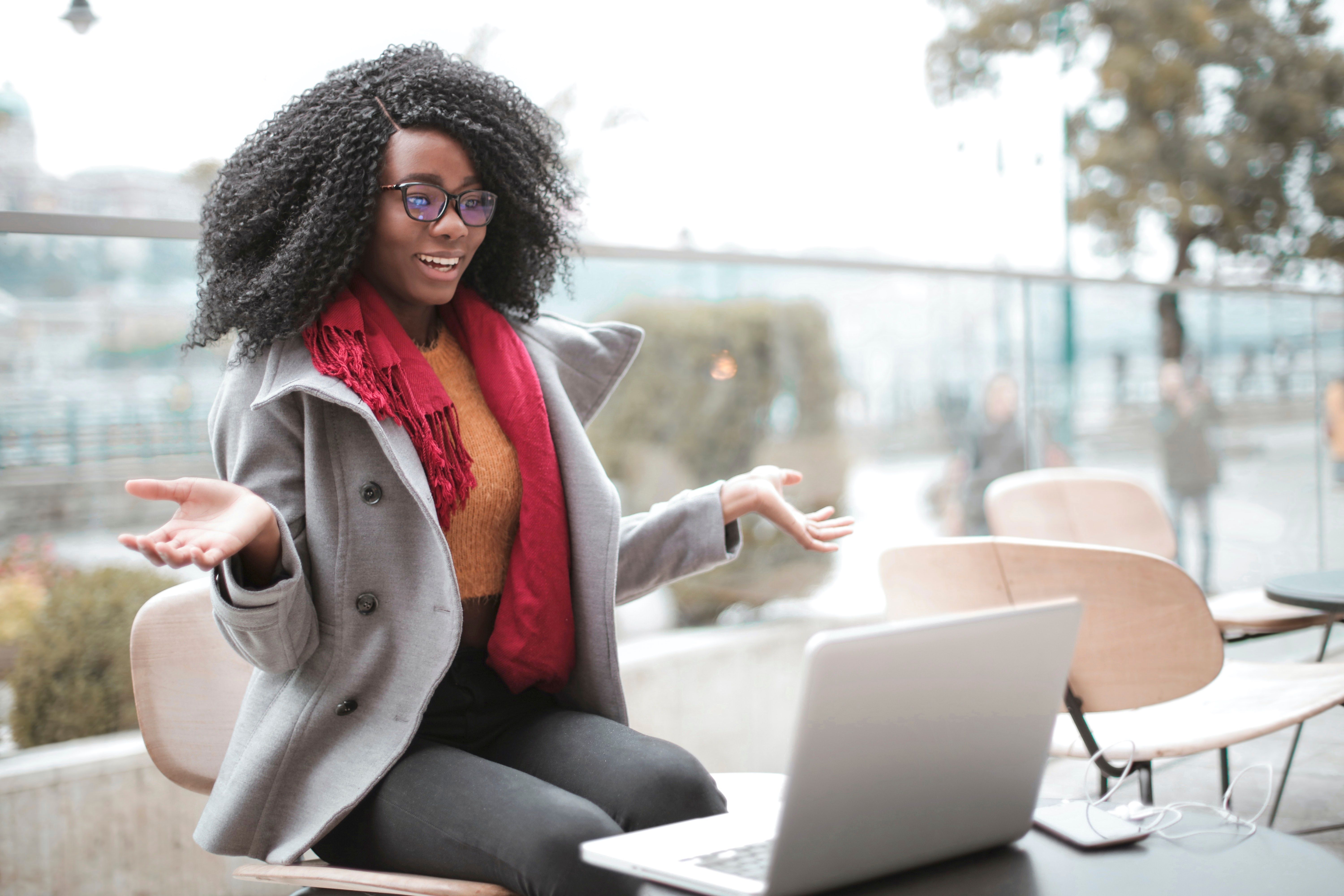 Woman with glasses and a red scarf gestures with her hands while looking at a laptop.