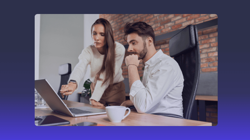 Two colleagues collaborate, discussing content on a laptop screen in a modern office.