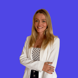 Smiling blonde woman in white blazer with arms crossed against a blue background.