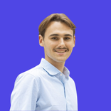 Young man with mustache and light blue shirt smiling against a purple background.