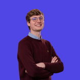 Smiling young man with glasses, wearing a maroon sweater, arms crossed, on a blue background.