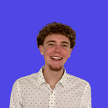 Smiling young man with curly brown hair wearing a white patterned shirt against a blue background.