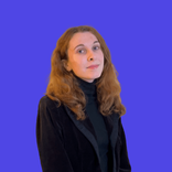 Young woman with wavy auburn hair wearing black, looking confidently at camera against purple background.